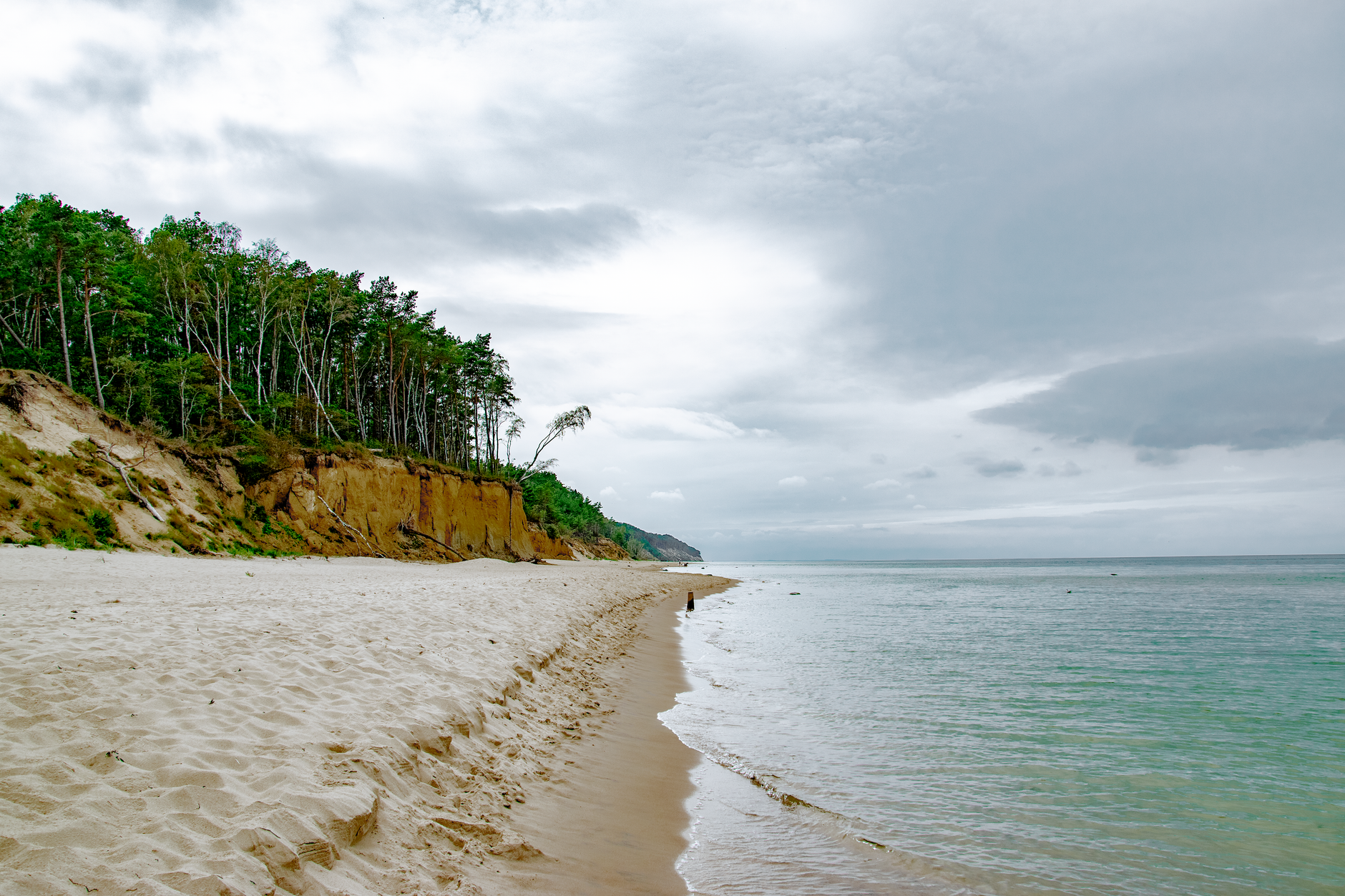 Serene beach with sandy shores, lush cliffs, and calm ocean in Świętouść, Poland under a cloudy sky.
