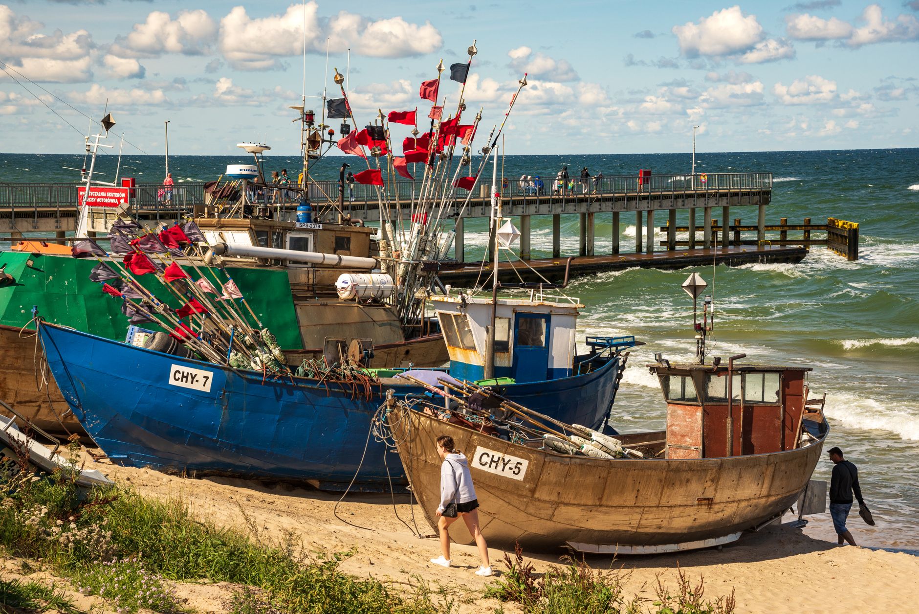 Colorful fishing boats docked on a sandy beach with a scenic ocean pier in the background on a bright sunny day.
