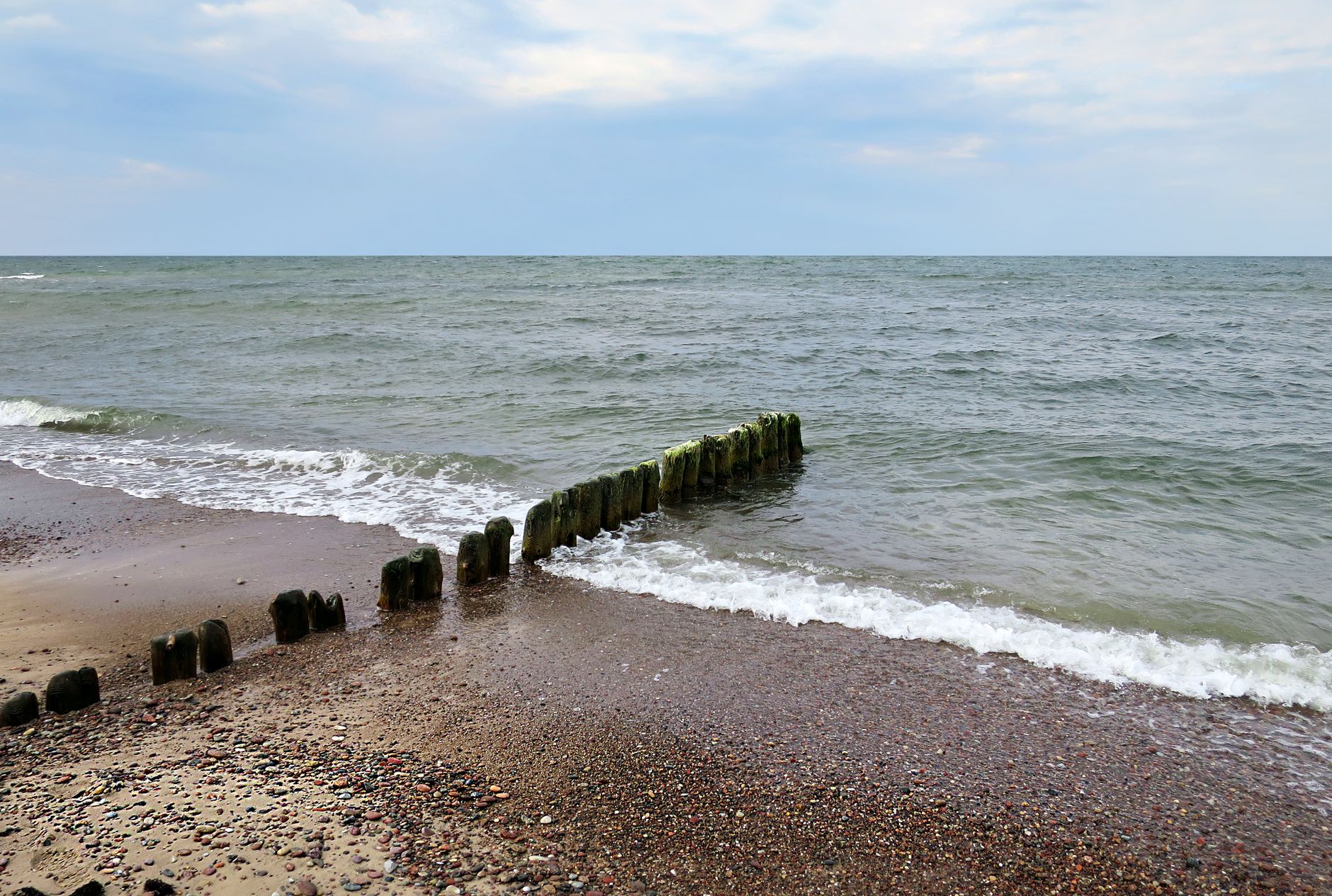 Serene view of Baltic Sea waves crashing against wooden breakwater on a pebbled beach.