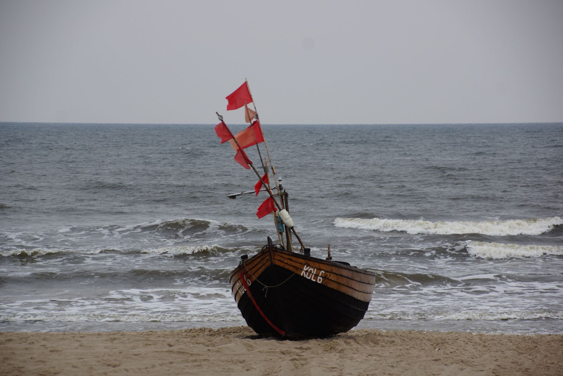 Wooden fishing boat with red flags on a beach by the ocean, under a cloudy sky.