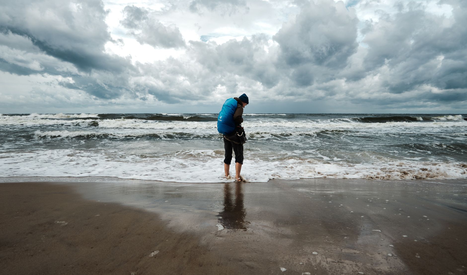 A person with a backpack stands on Kopań Beach in Poland, facing rough ocean waves under a dramatic sky.