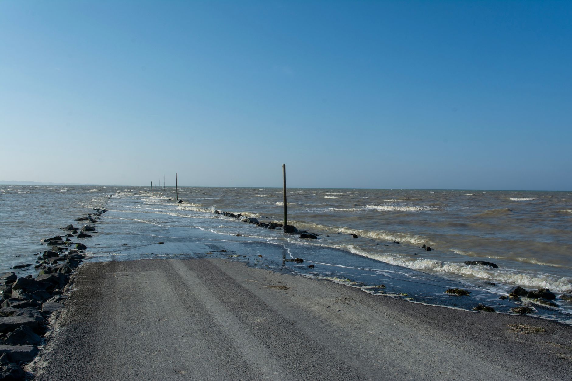 A flooded coastal road surrounded by the sea and horizon under a clear blue sky.