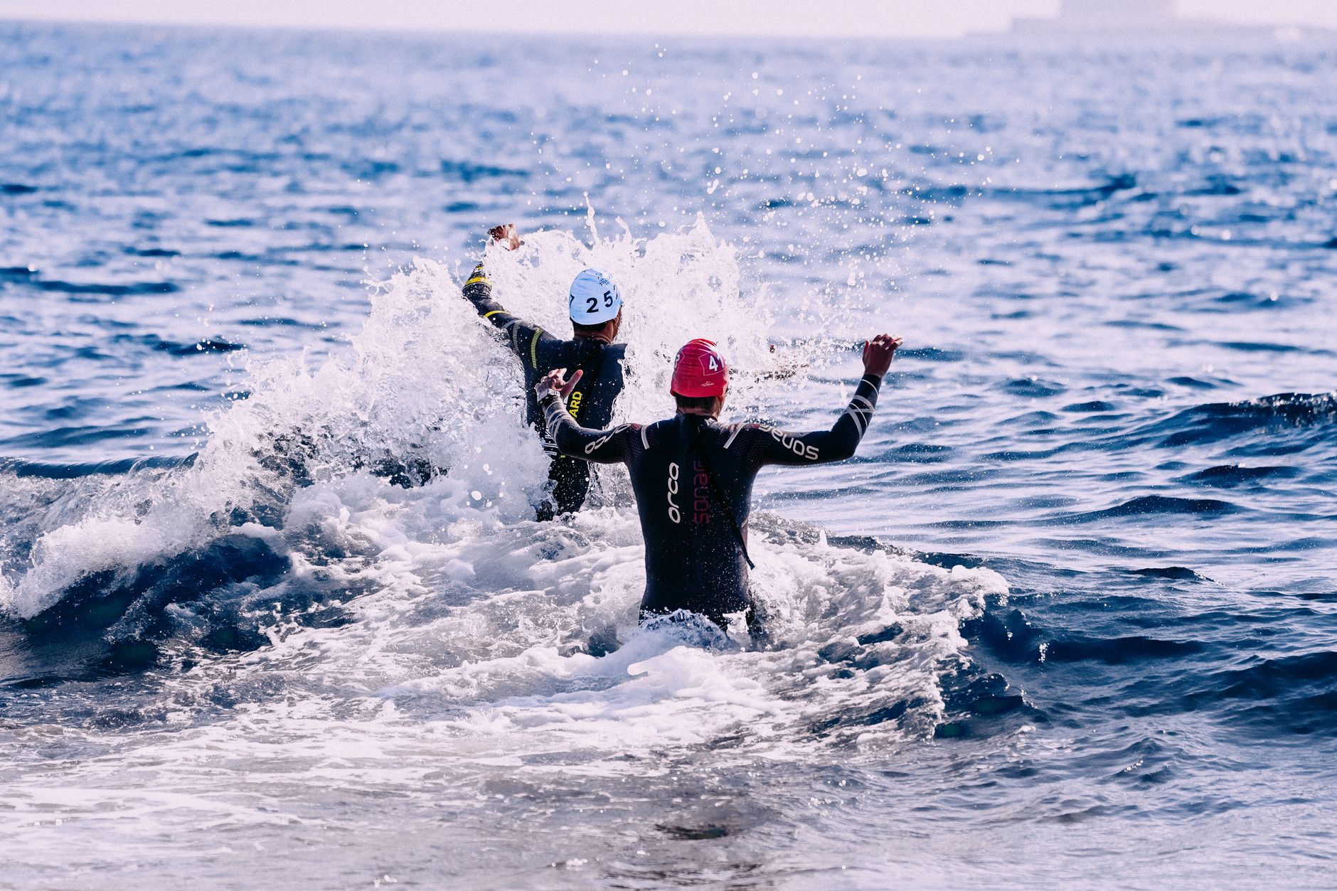 Back view of faceless sportsmen in wetsuits and caps coming into vibrant dark blue wavy ocean in daytime