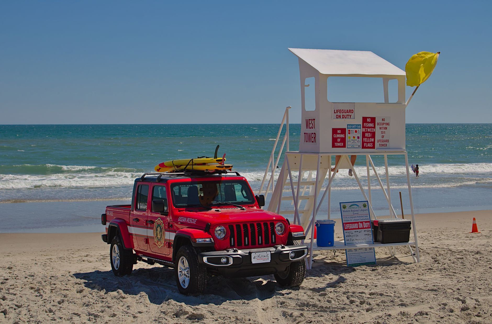 Red lifeguard vehicle and tower on a sandy beach with the ocean in the background, under a clear blue sky.