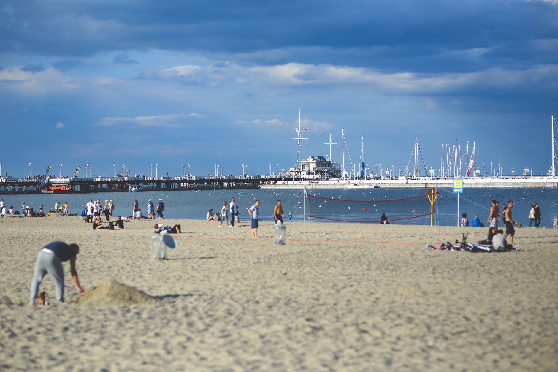 A lively beach scene featuring people playing volleyball, relaxing, with boats in the background under a blue sky.