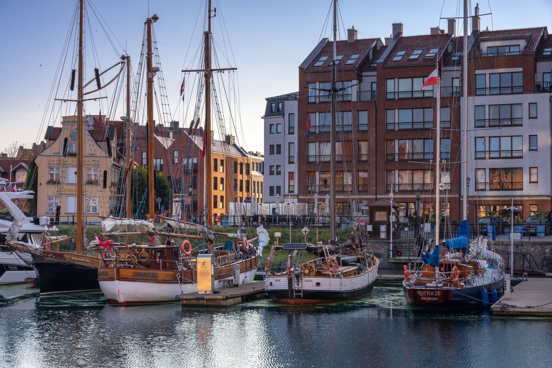 Scenic view of a marina in Gdańsk featuring classic sailing boats and historic buildings at sunset.