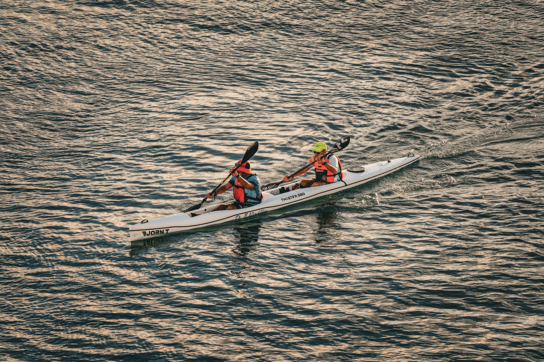 Two adults kayaking on a calm body of water, wearing life vests and helmets during the day.