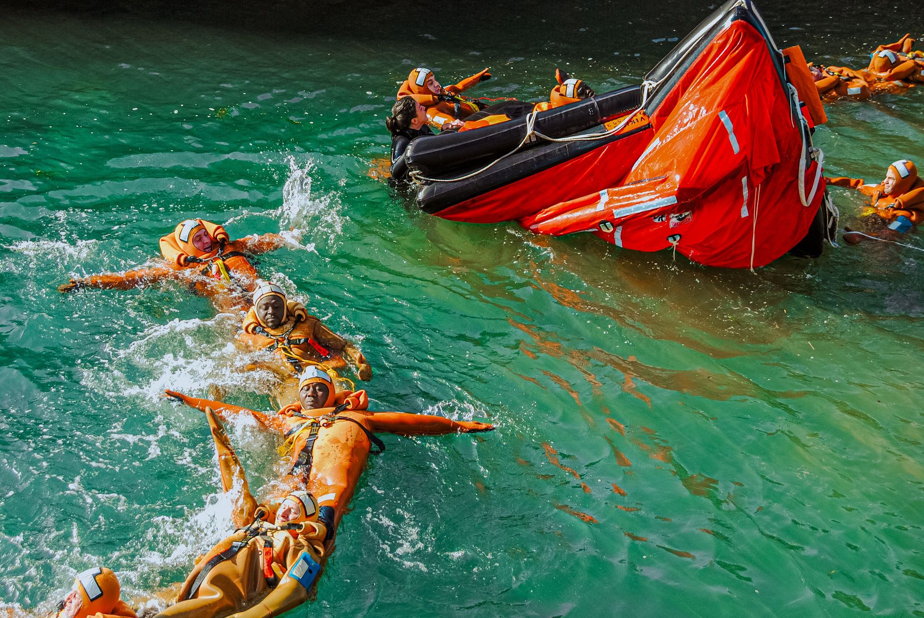 Group of adults practicing lifeboat safety drill in the water wearing life jackets.