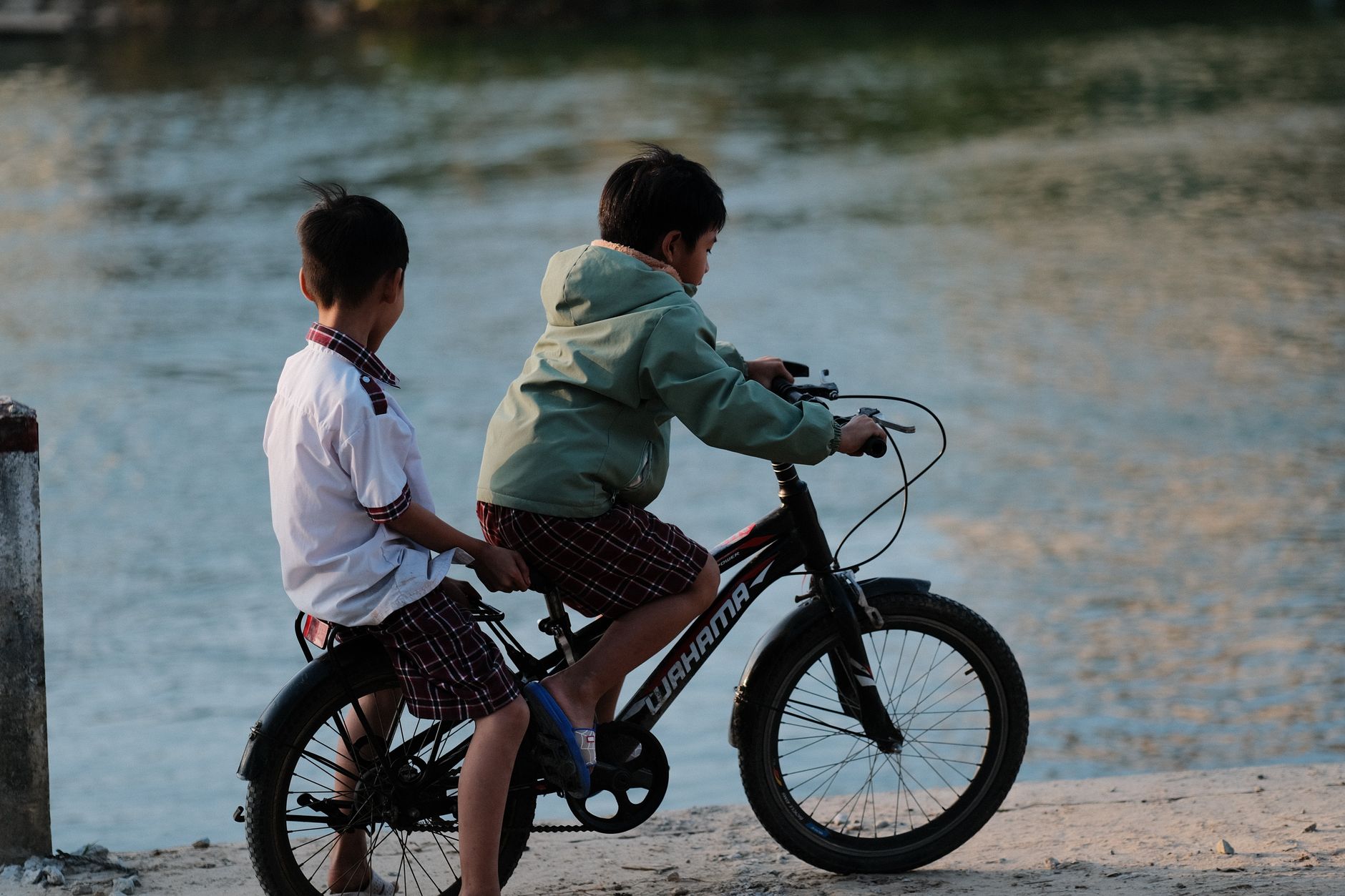 Two boys enjoying a ride on a bicycle by the waterside during the day.