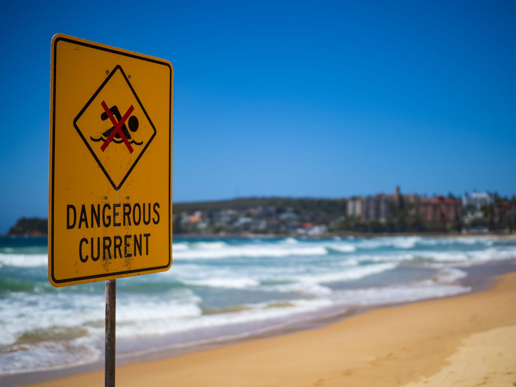 Dangerous current warning sign on Manly Beach in Australia, with blue sky and ocean waves.