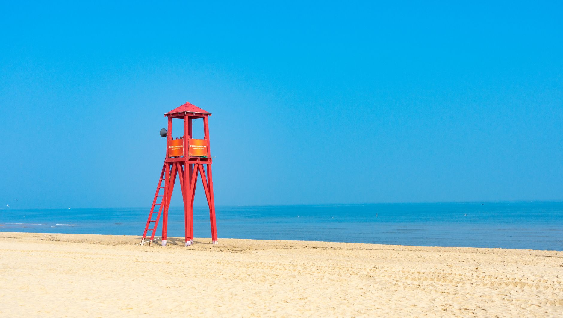 Red lifeguard tower stands on a pristine sandy beach under a clear blue sky.