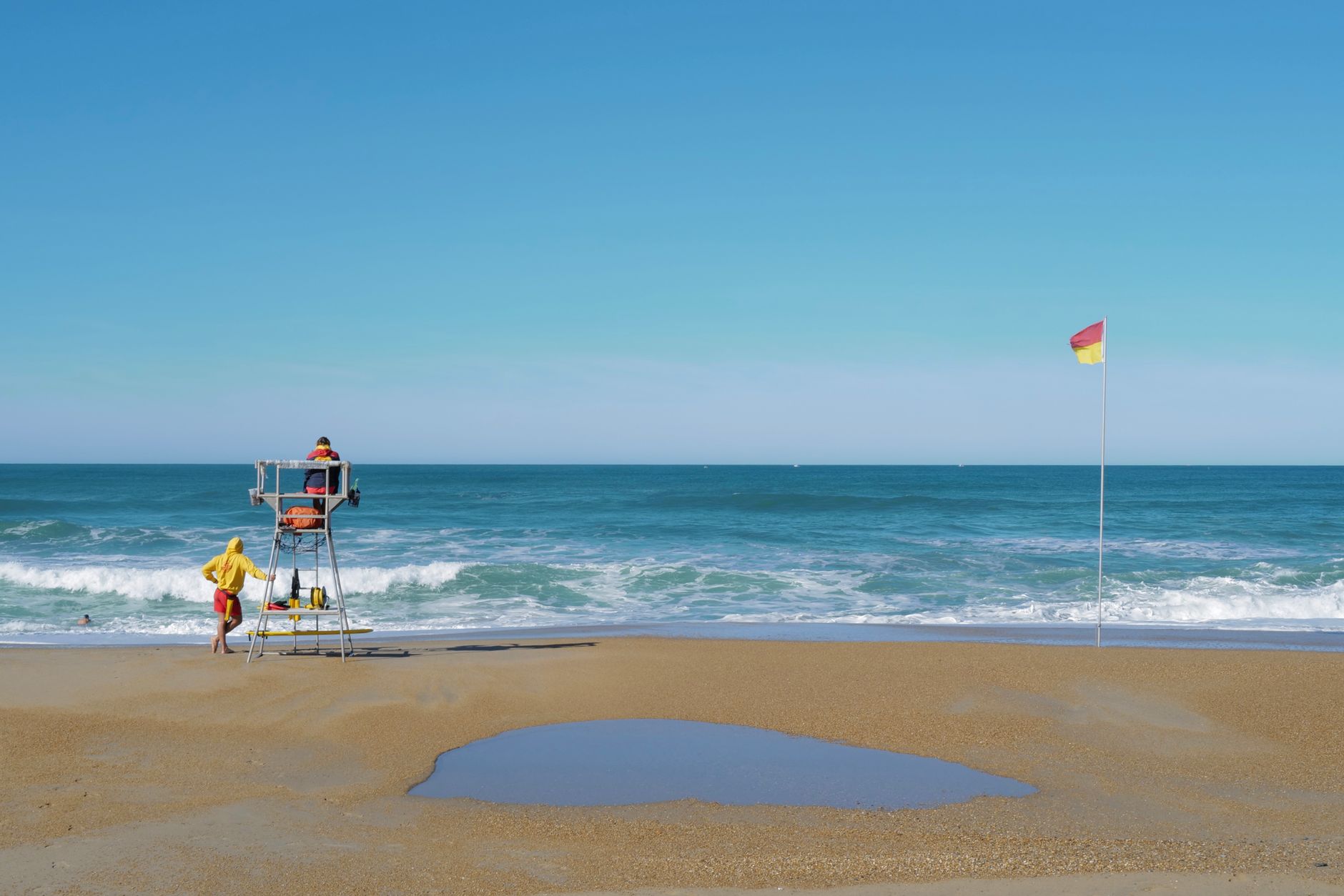 Serene beach scene with lifeguards, waves, and safety flag under a clear sky.