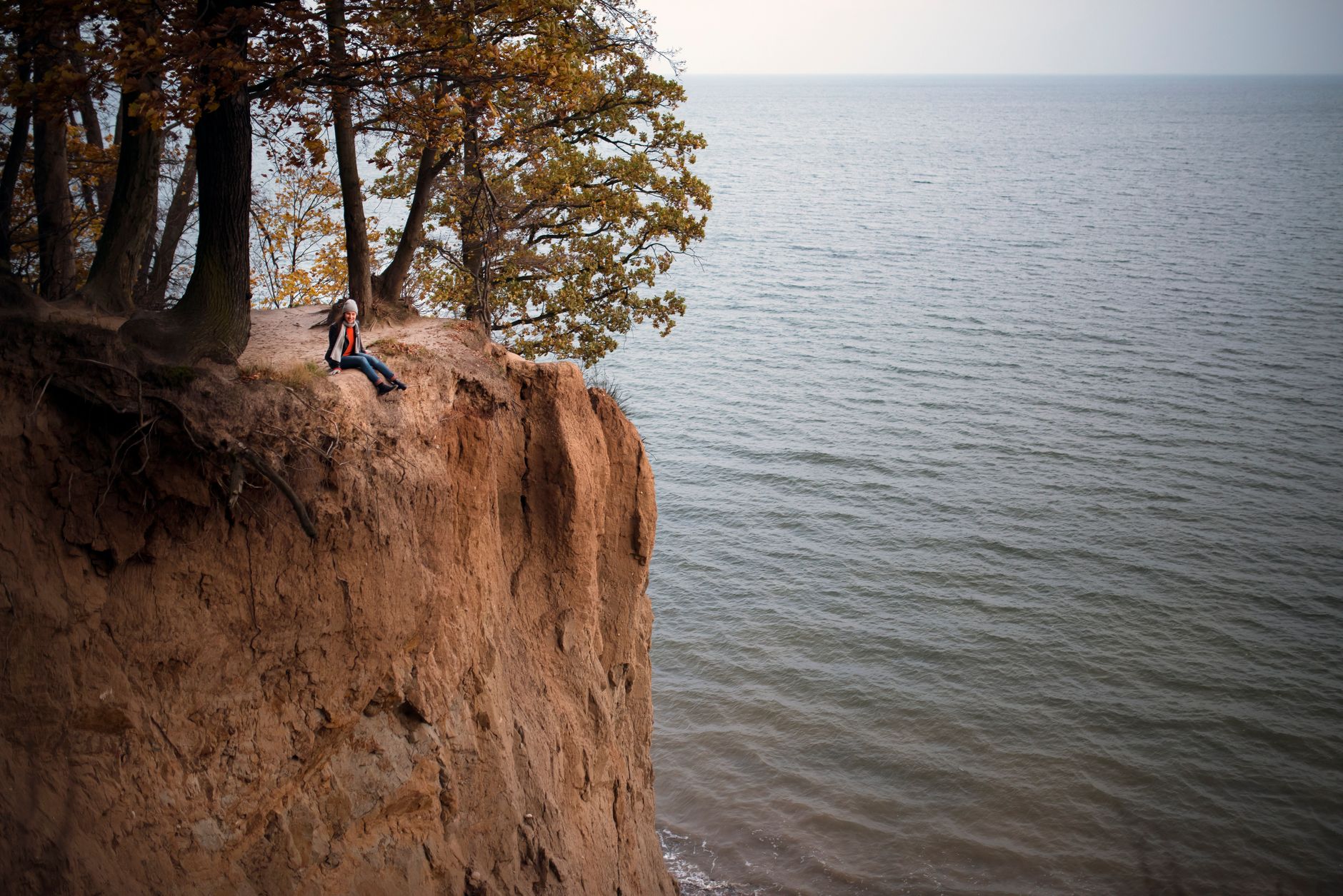 A woman sits on a cliff edge under trees, overlooking the vast sea in Gdynia, Poland.