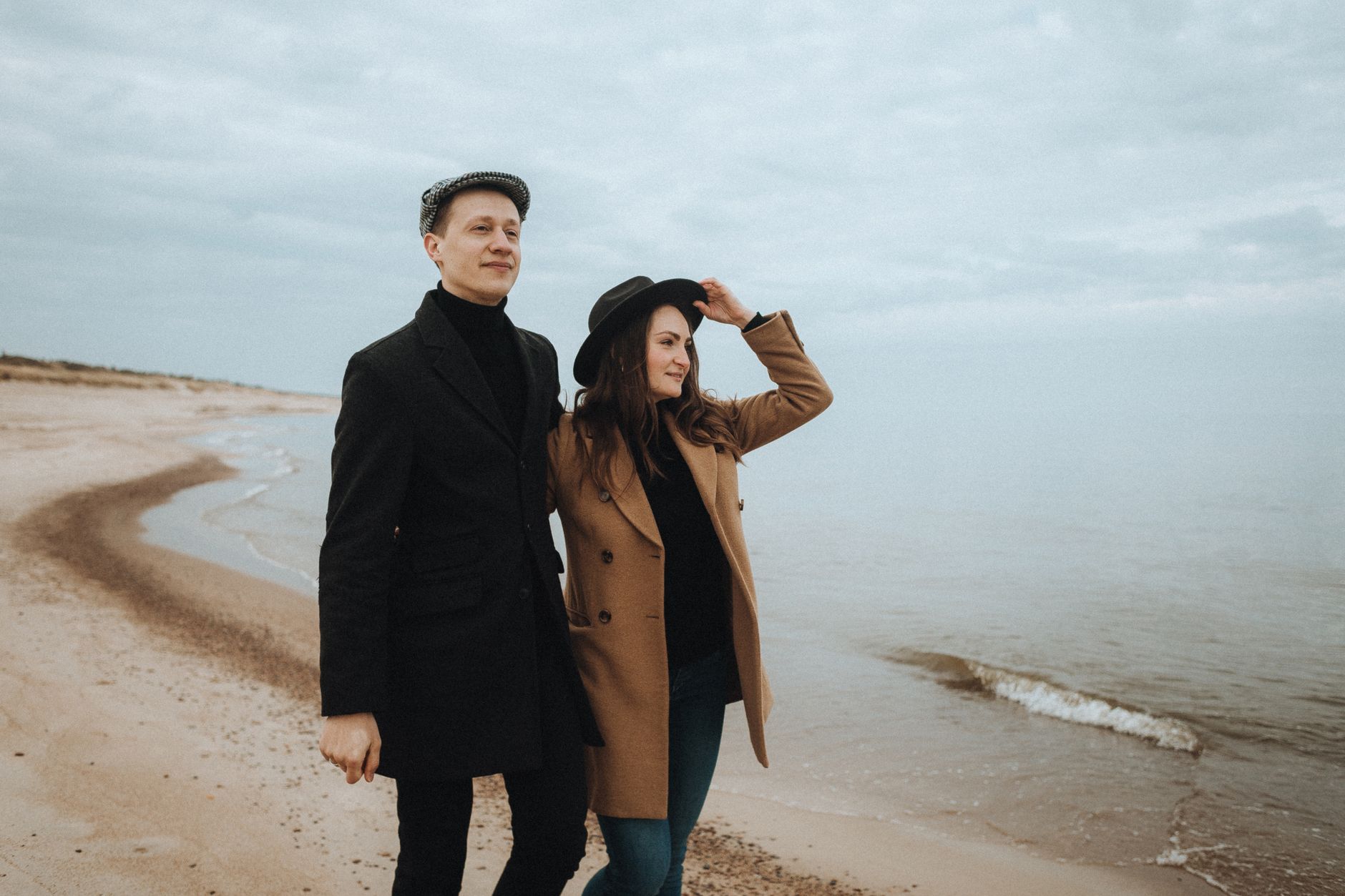 Couple in coats walking together on a serene beach shore under cloudy skies.
