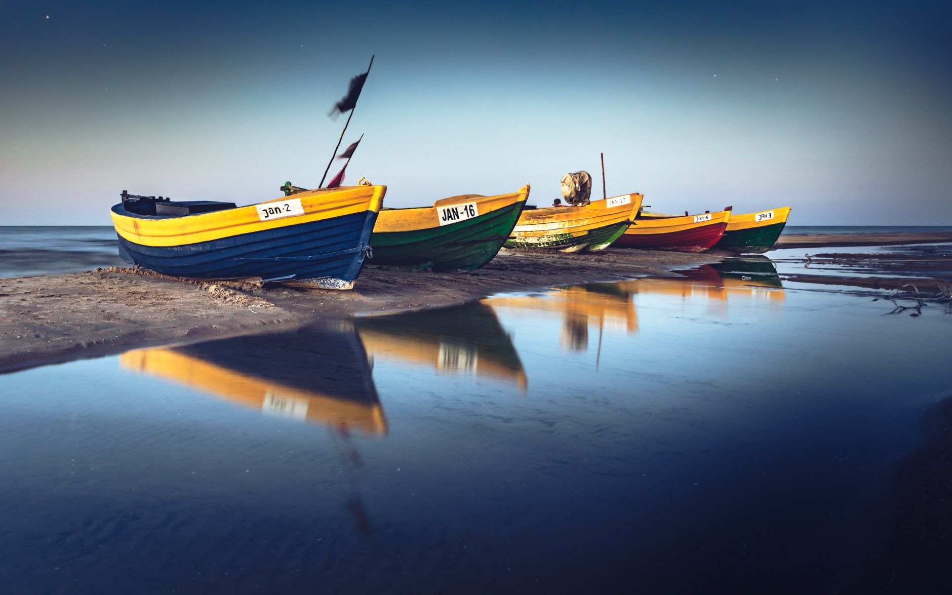 Vibrant fishing boats reflect on the tranquil shore of Jantar beach, Poland at sunset.
