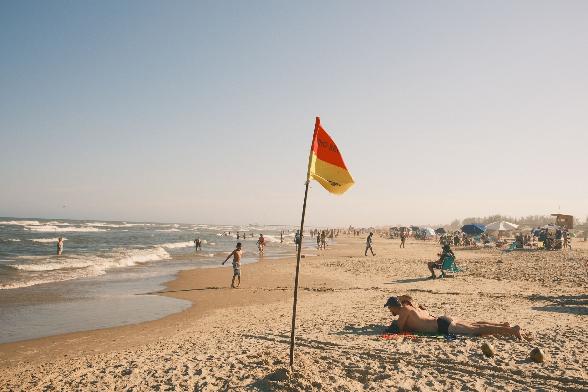 A vibrant beach scene with people enjoying the sun and surf, featuring a prominent lifeguard flag.