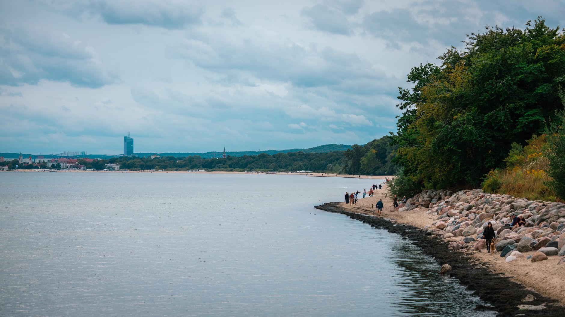Tranquil beach scene with people walking, trees, and a distant city skyline under a cloudy sky.