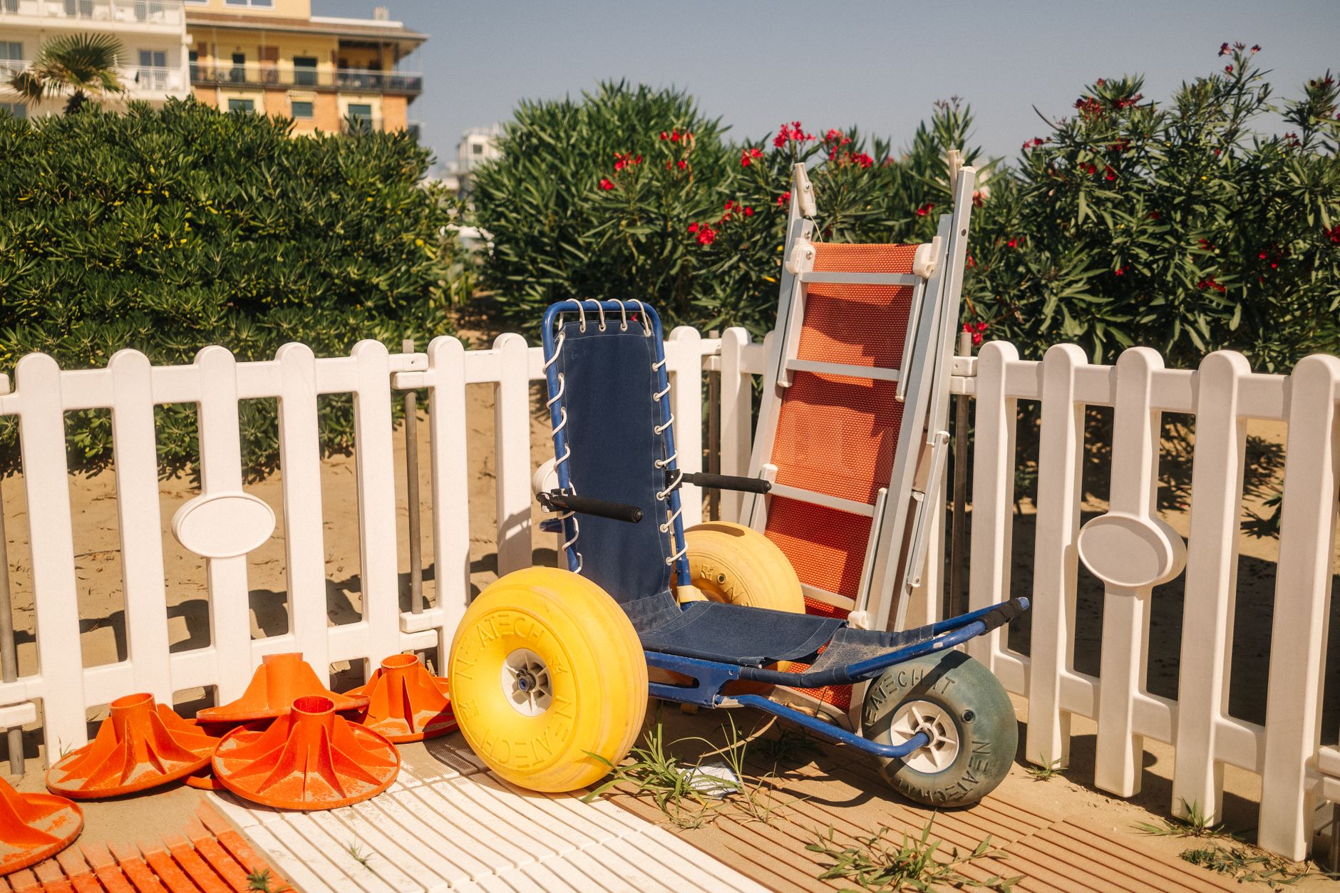 Beach wheelchair next to lifeguard equipment by a white fence on a sunny day.