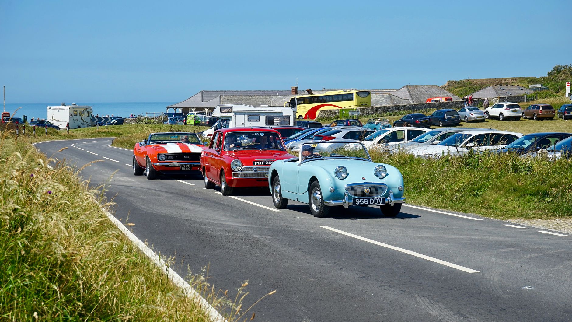 Classic cars cruise along a picturesque coastal road in Eastbourne, UK, on a sunny day.