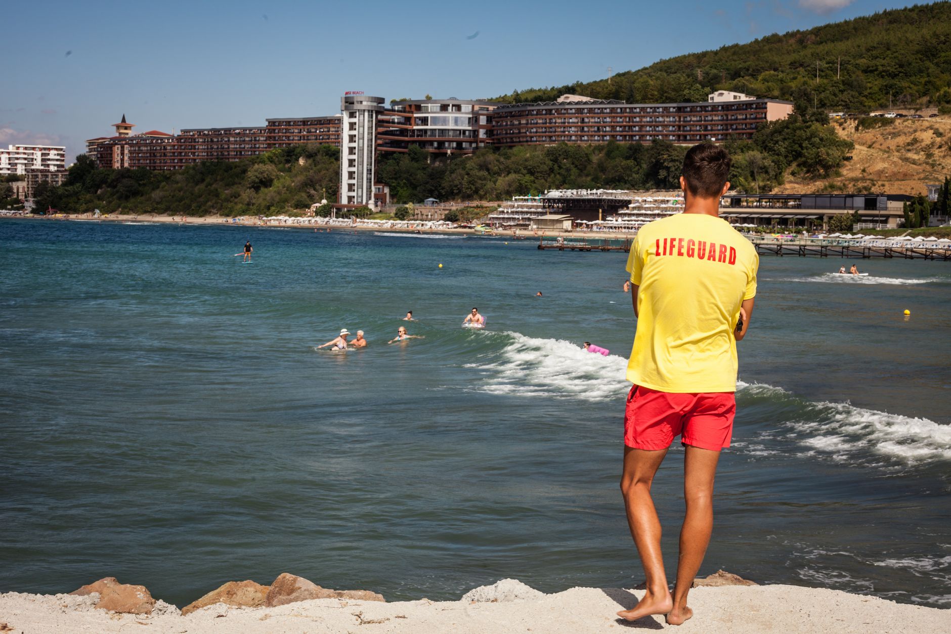 Lifeguard in yellow shirt watches over swimmers near beachfront hotels.