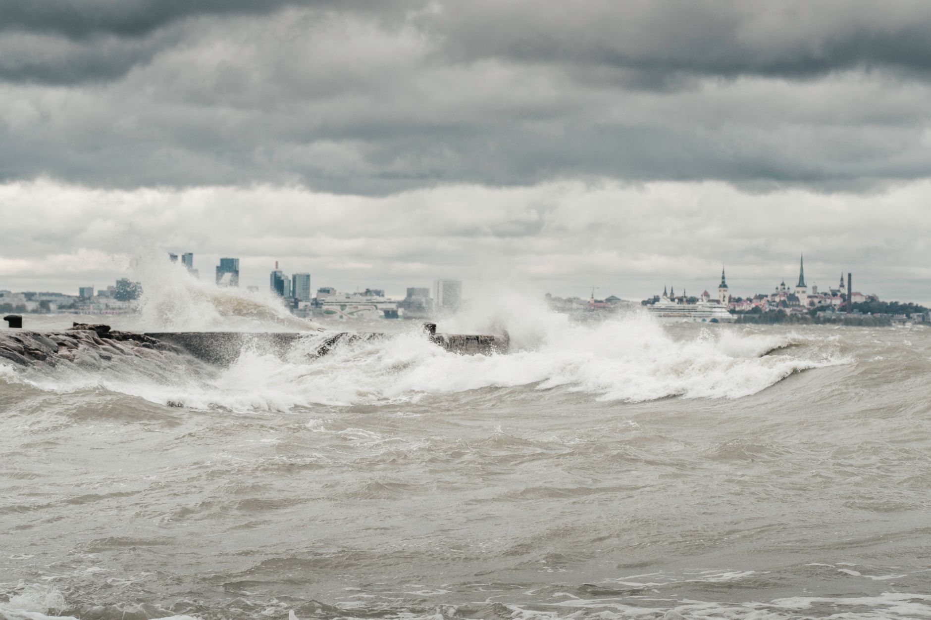 Powerful waves crash against a seawall with Tallinn's skyline in the distance, under a moody sky.