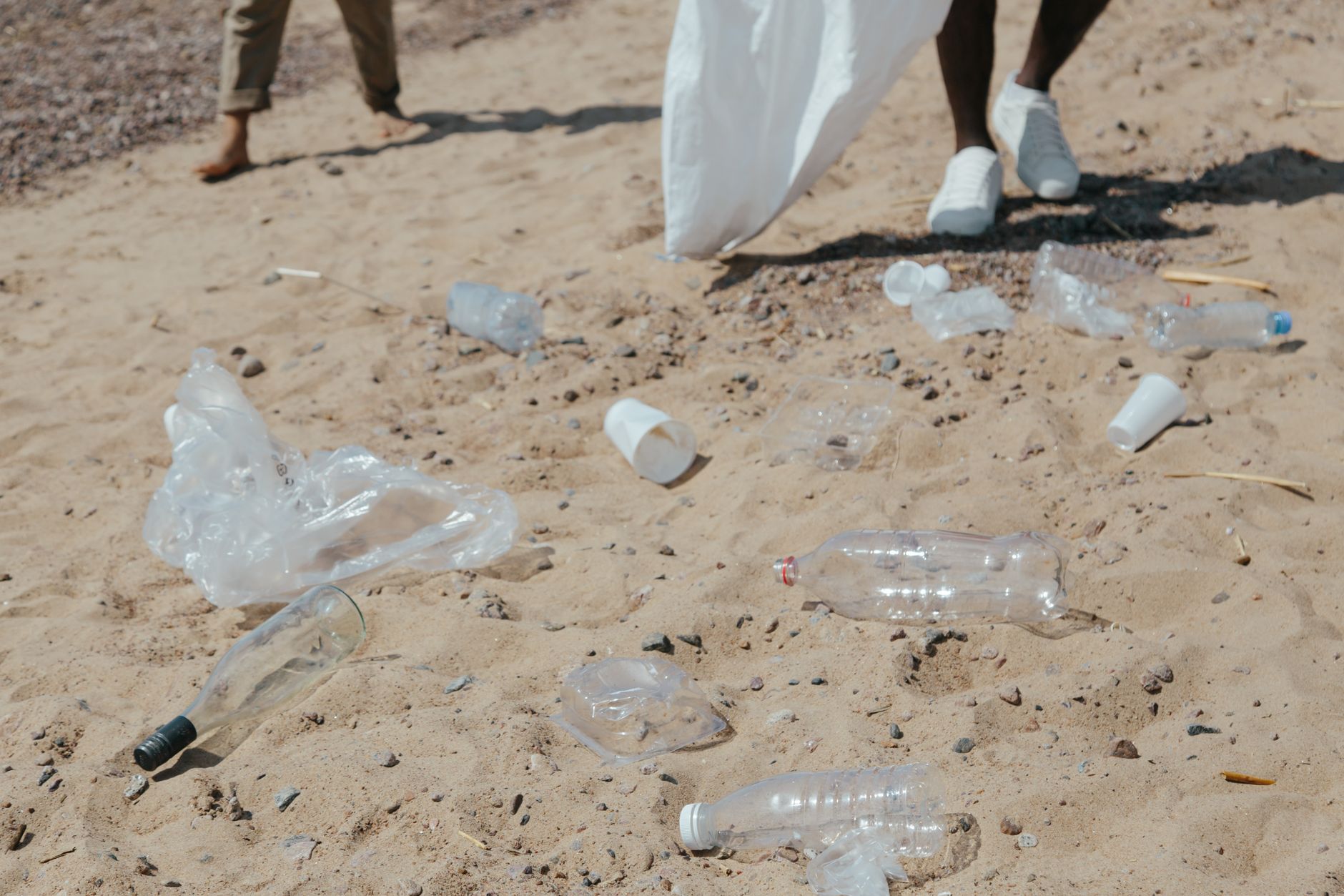 Plastic waste scattered on a sandy beach with people collecting it for cleanup.
