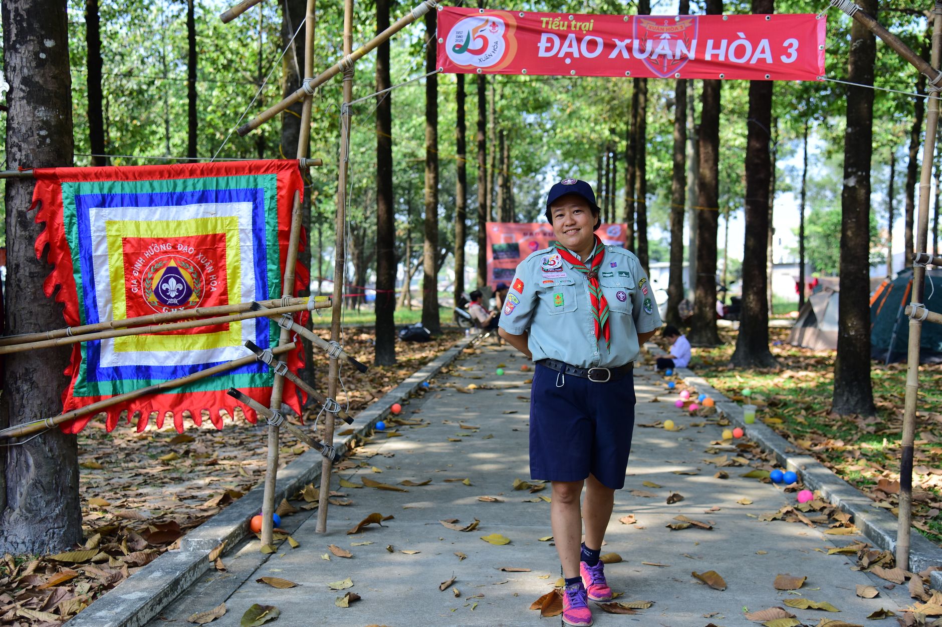 Scout leader in uniform at an outdoor event in a wooded park area with decorations.