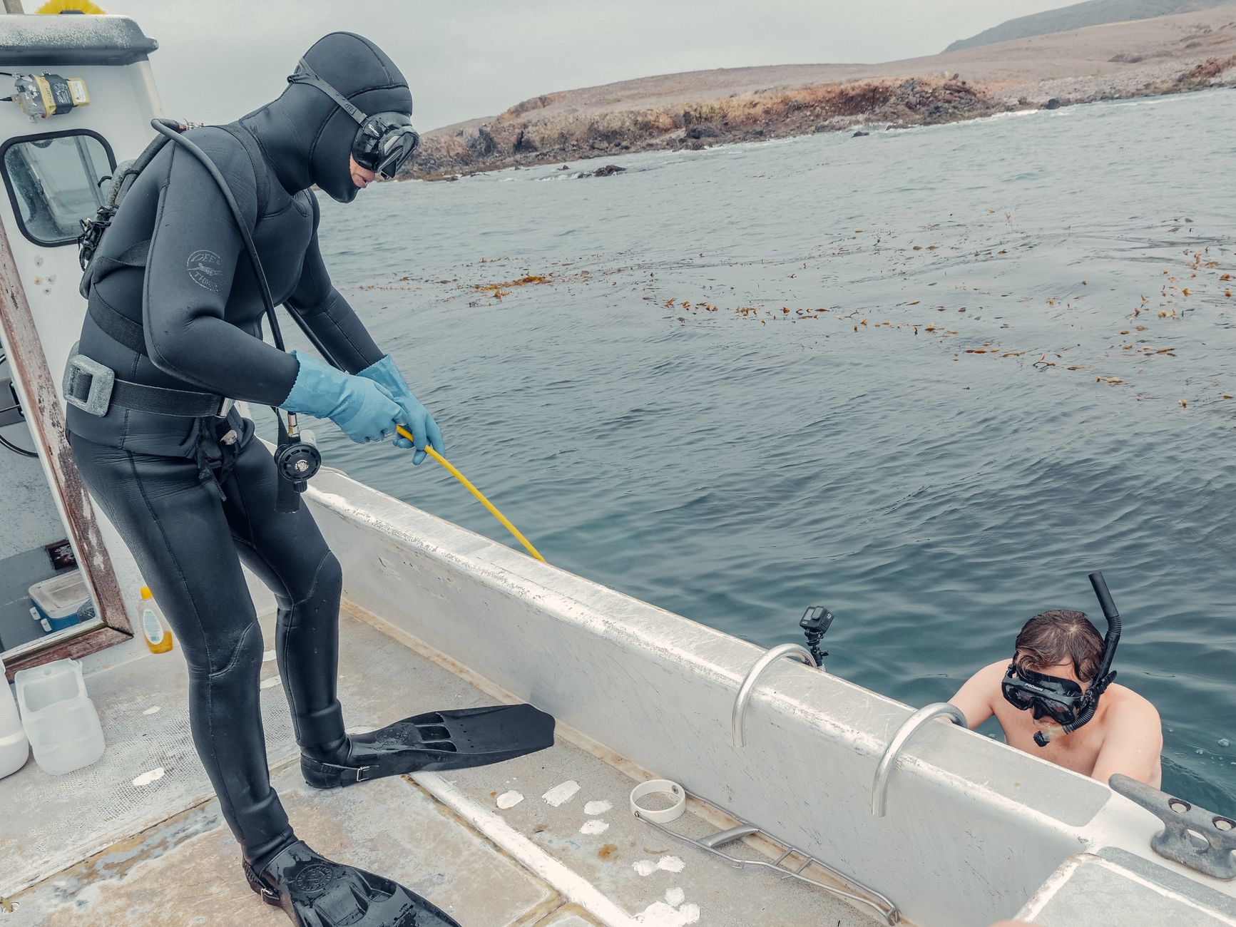 Two divers prepare on a boat, one in a wetsuit standing with gear, another in water holding the boat.
