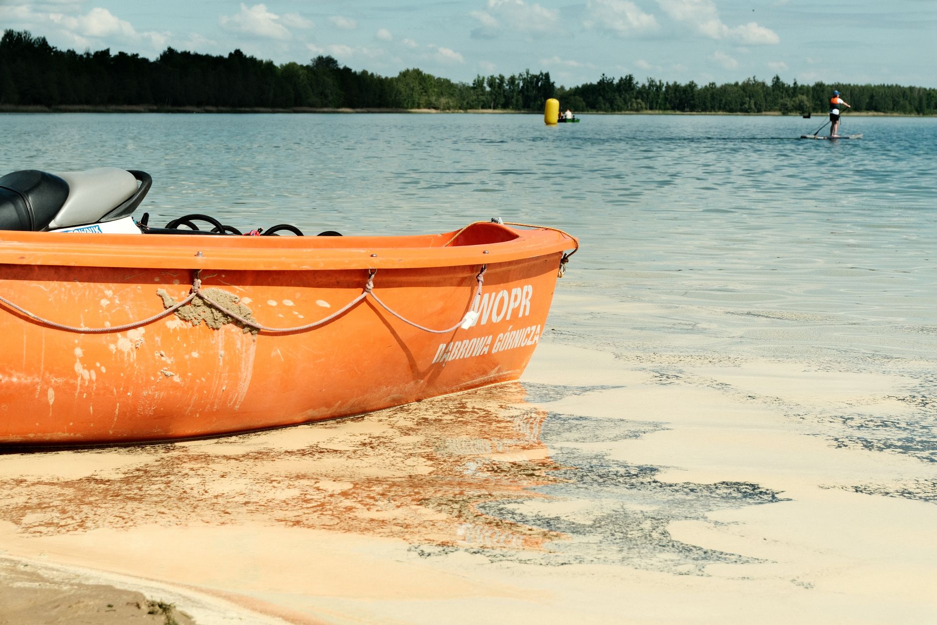 Sunny day at a Polish lake with an orange lifeboat and paddleboarder in view.