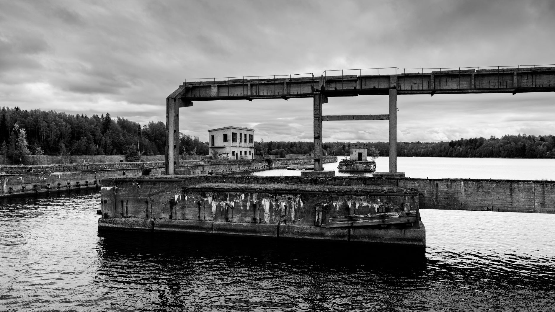 Black and white view of an abandoned industrial facility by a tranquil lake under a cloudy sky.