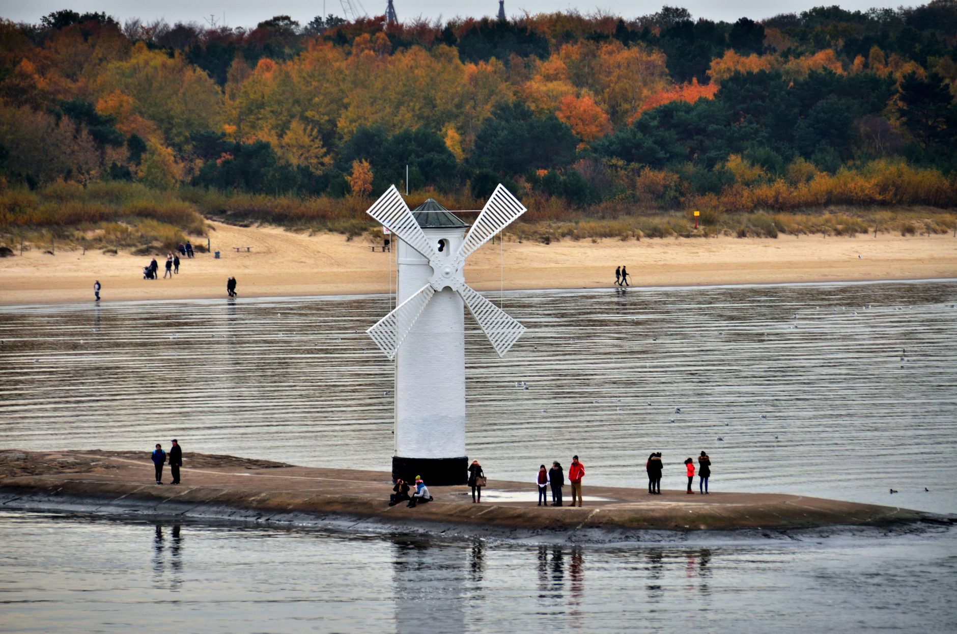 Scenic view of the iconic Stawa Młyny beacon with autumn foliage and beachgoers in Świnoujście, Poland.
