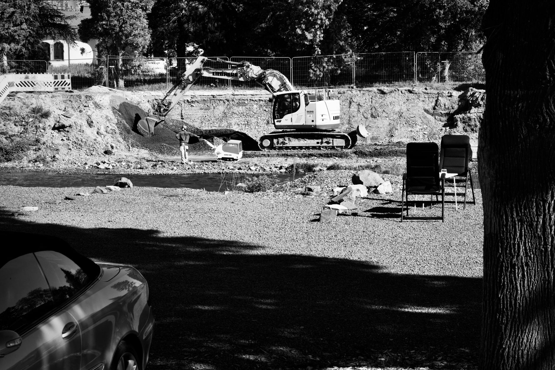 Monochrome image of an excavator at a construction site next to a river with folding chairs nearby.