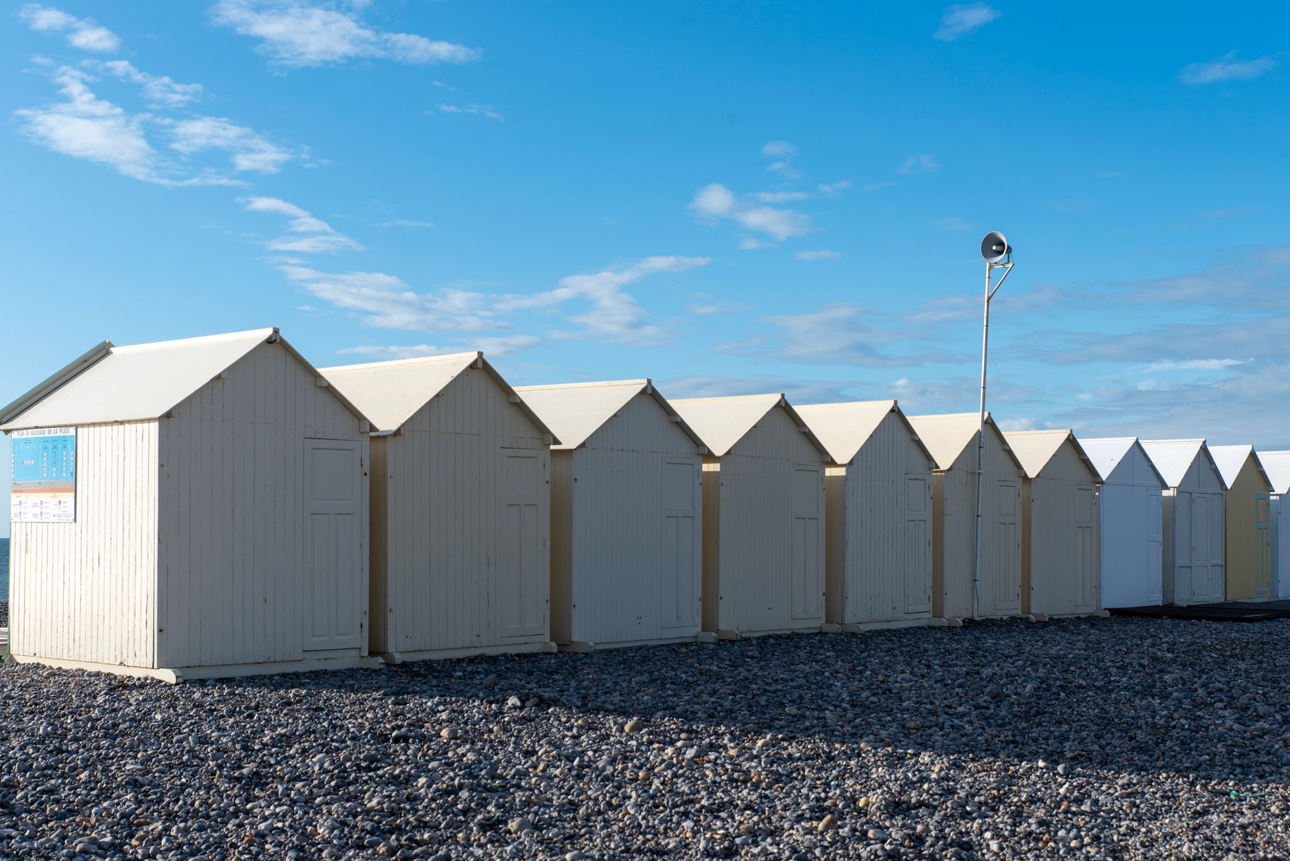 White beach huts lined up on a pebbled shore under a bright blue sky, captured in daylight.