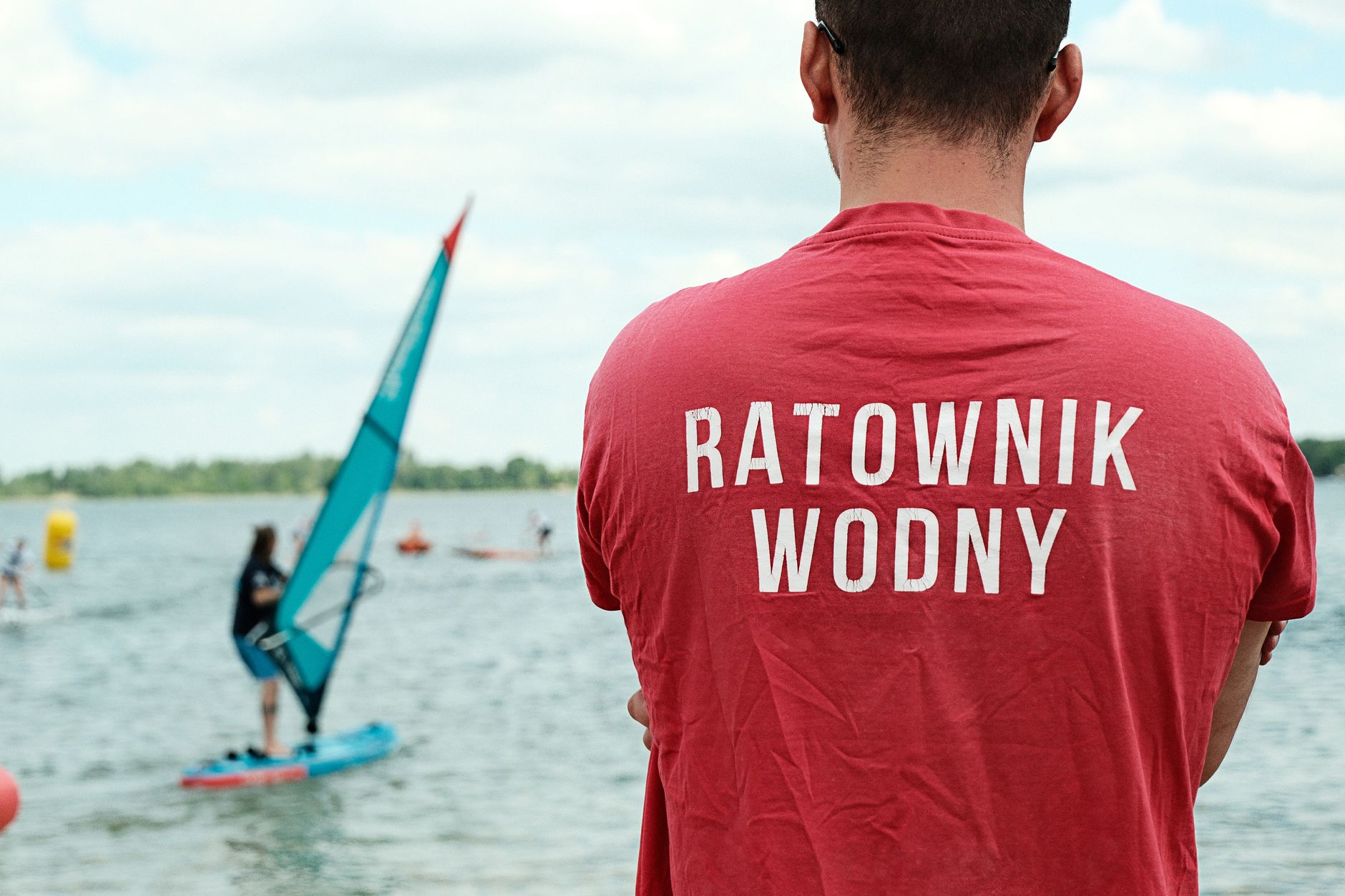 Lifeguard supervising a paddleboarder on a Polish lake, under a partly cloudy sky.